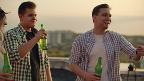 Young-men-and-women-have-enjoyable-time-on-the-roof.-They-smile-and-communicate-with-each-other.-They-drink-beer-from-green-bottels-and-have-fun-in-the-plaid-shirts.